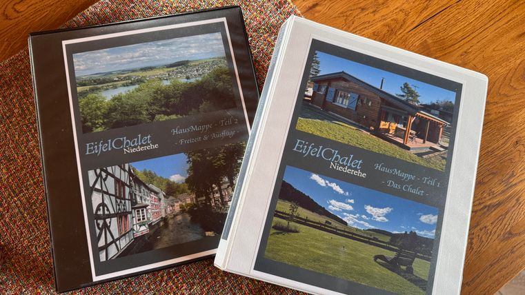 Zwei Ordner mit Fotos und Informationen über das EifelChalet. Einer zeigt eine Fachwerkhausansicht und der andere das Chalet in der Natur.