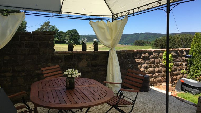 Un espace extérieur confortable avec une table ronde en bois et des chaises. En arrière-plan, on aperçoit un joli paysage avec des collines.