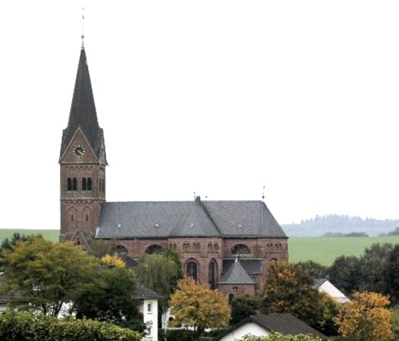 Church with a high tower in a rural setting, surrounded by trees and houses. The sky is cloudy., © Touristik GmbH Gerolsteiner Land