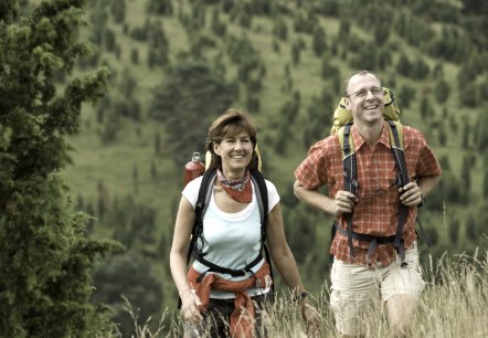 Een vrouw en een man in wandelkleding en met rugzakken lopen over een weide met hoog gras. Op de achtergrond jeneverbesvelden.