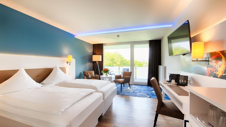 Ein modernes Hotelzimmer mit zwei Betten, einem Schreibtisch und einem Fenster mit Blick auf die Natur. Die Wände sind blau und die Einrichtung ist hell und freundlich.