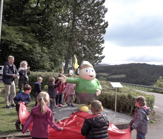 Statue von Maskottchen Willi Basalt mit gr&uuml;nem Shirt, grauer Hose und gelben Schuhen zeigt nach links. Um die Figur herum einige Kinder im Kreis.