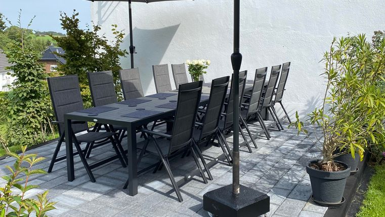 Ein Außensitzbereich mit einem großen Tisch und schwarzen Stühlen. Zwei Sonnenschirme bieten Schatten auf der gepflasterten Terrasse.