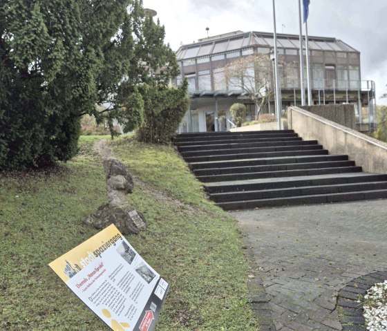 Modern gebouw met glazen gevel, trappen en vlaggen. Op de voorgrond een bord op een weiland omringd door bomen., © Clara Zins-Grohé
