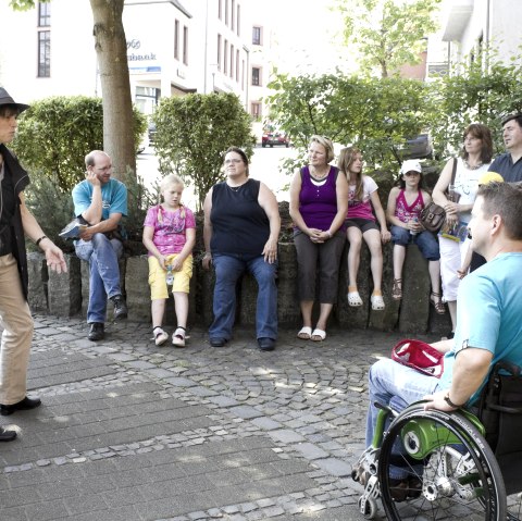 Eine Gruppe von etwa 11 Personen bestehend aus Erwachsenen und Kindern sitzt nebeneinander in einem Kreis auf einer Mauer neben einer Straße. Vor der Gruppe steht eine Gruppenführerin und erklärt etwas.