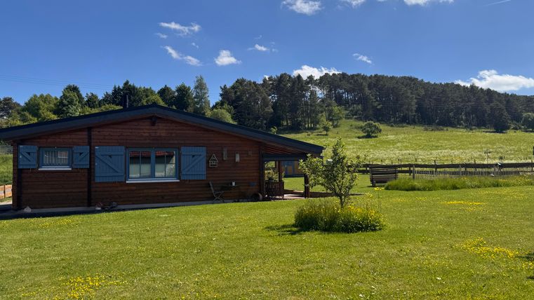 Ein Holzhaus umgeben von grünem Rasen und Bäumen. Der Himmel ist blau mit wenigen Wolken.
