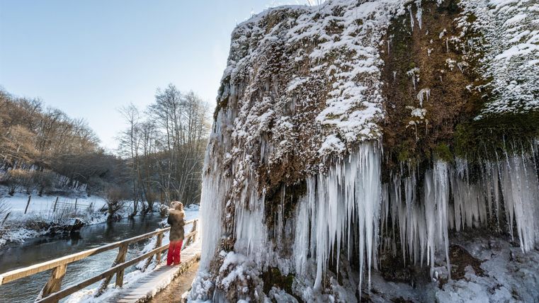 Gefrorener Nohner Wasserfall mit Eiszapfen, eine Person auf einem Steg, winterliche Landschaft mit Schnee und kahlen Bäumen.