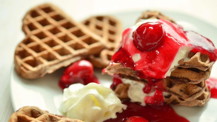 Herzförmige Waffeln liegen nebeneinander und gestapelt auf einem Teller mit Sahne und Kirschsoße darauf.