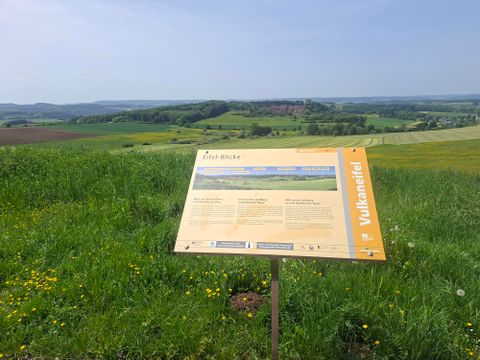 Ein Informationsschild steht auf einer grünen Wiese mit Blick auf die hügelige Landschaft. Im Hintergrund sind Wälder und Felder zu sehen.