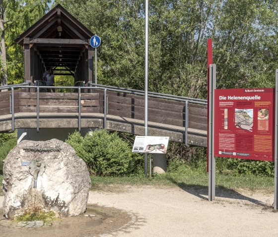 Overdekte houten brug en informatiebord naar de bron Helenenquelle in het Kyllpark Gerolstein, omgeven door bomen en een grote steen met een kraantje., © Jochen Hank