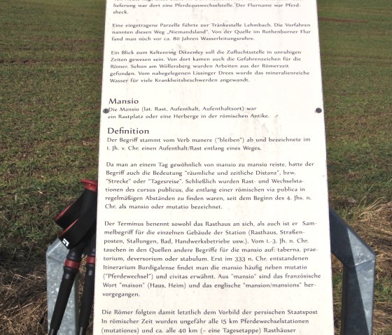 Information board about a Roman horse changing station, explaining historical details and terms. Meadow and trees in the background, walking sticks in front., © Touristik GmbH Gerolsteiner Land, Ute Klinkhammer