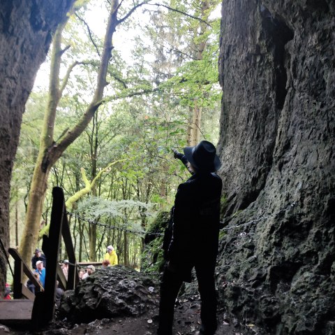 Stone entrance to a cave in which a person is standing and pointing a gun at the forest in front of the cave. A staircase leads up to the cave and several people are standing at the top of the stairs.