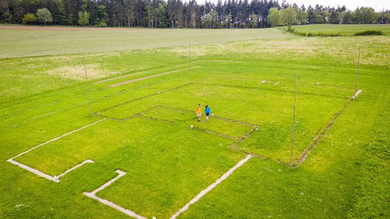 Luftaufnahme von zwei Personen auf einem Feld mit markierten Grundrissen einer römischen Villa.