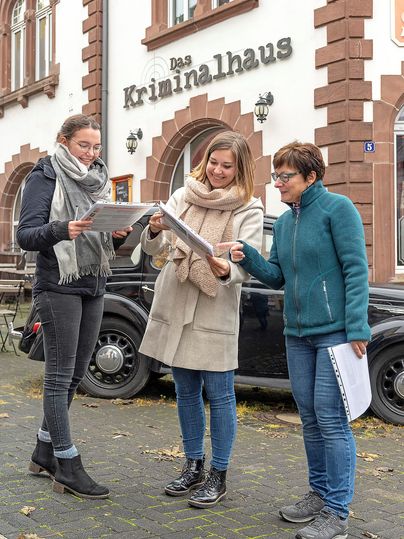 Drei Frauen stehen lachend vor dem Kriminalhaus Hillesheim und betrachten Dokumente. Im Hintergrund ist ein altes Auto zu sehen.