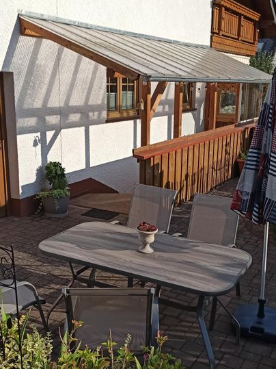 Un coin salon confortable à l'extérieur avec une table et des chaises. À l'arrière-plan, on voit une maison lumineuse et des plantes.