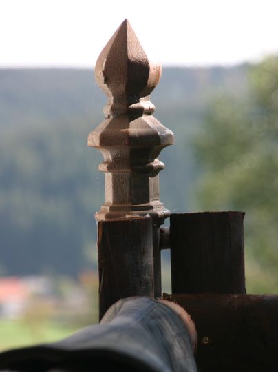 Een decoratieve houten paal met een spitse vorm op de voorgrond. Op de achtergrond zijn vage groene landschappen te zien.