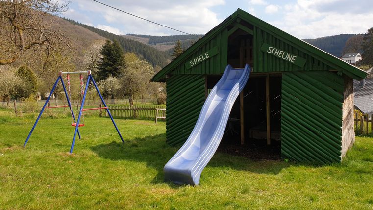 Een speelplaats met een glijbaan en een schommelpaal staat op een groen veld. Op de achtergrond zijn zachte heuvels en een blauwe lucht te zien.