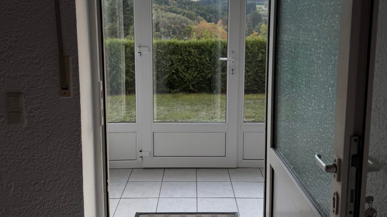 Une entrée lumineuse avec un sol en carrelage et un paillasson. À travers de grandes fenêtres, vue sur un paysage verdoyant.