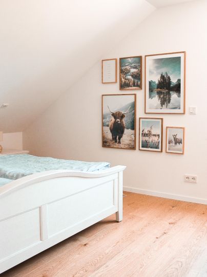 Ein gemütliches Schlafzimmer mit einem weißen Bett und einer Holzboden. An der Wand hängen mehrere Bilder in Rahmen, die Landschaften und Tiere zeigen.