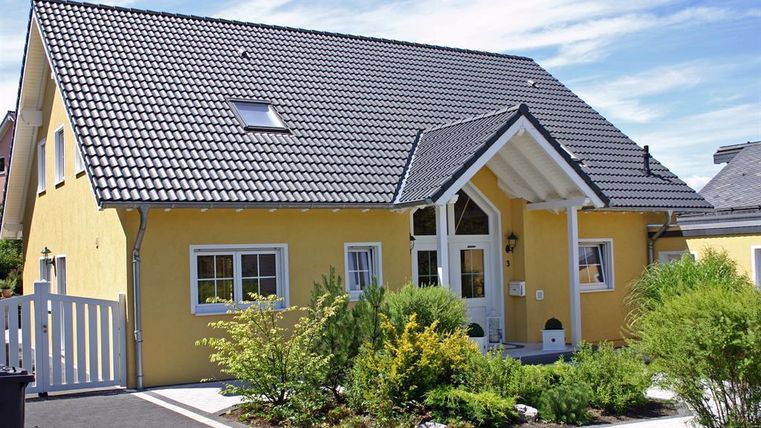 Een modern, geel eengezinswoning met een schuin dak. De tuin is groen en goed onderhouden, met veel planten en een toegangspoort.