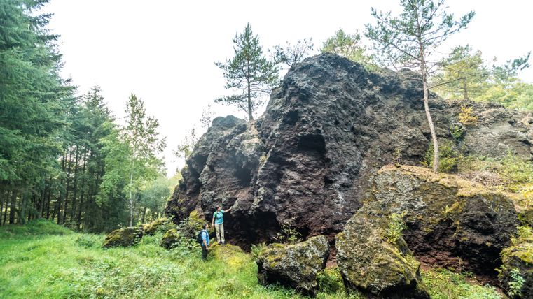 Zwei Personen stehen vor einem großen, dunklen Felsen in einem Waldgebiet.