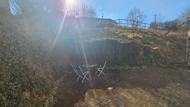 Un espace extérieur ensoleillé avec une petite table et deux chaises. L'endroit est entouré d'une haie verte et possède un sol pavé.