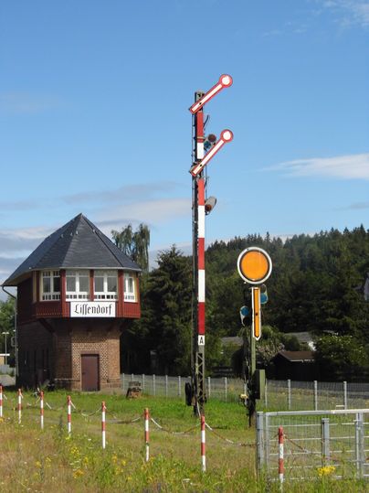 Ein altes Stellwerkgebäude mit der Aufschrift 'Lissendorf' und ein mechanisches Eisenbahnsignal vor blauem Himmel.