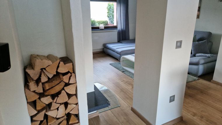Ein modernes Wohnzimmer mit Holzregal und gemütlicher Sitzecke. Große Fenster sorgen für viel Tageslicht.