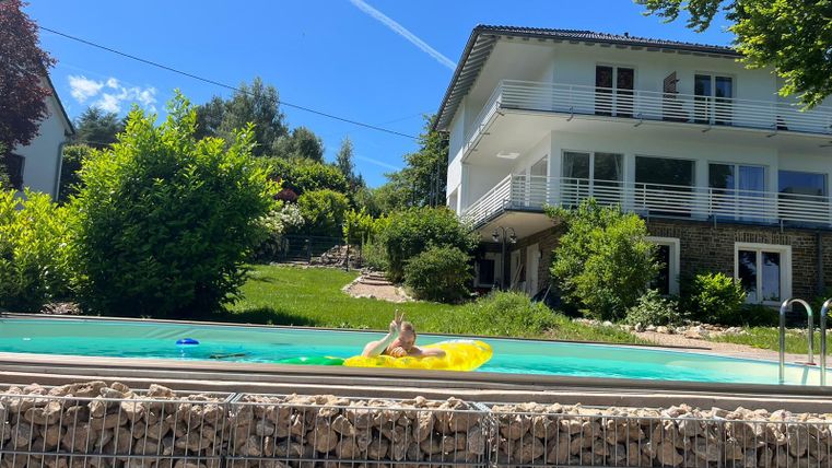 Ein ruhiger Garten mit einem Schwimmbad und üppigem Grün. Eine Person entspannt sich auf einer Luftmatratze im Wasser.