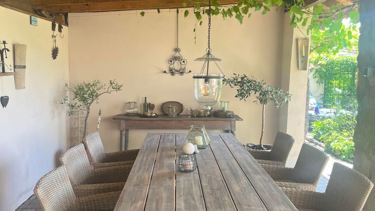Un coin repas confortable en plein air avec une longue table en bois et des chaises en osier. Au-dessus de la table, une lampe décorative pend, et des plantes créent une atmosphère agréable.