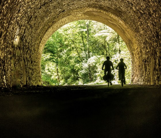 Zwei Radfahrer durchqueren einen Tunnel auf dem Ahr-Radweg. Am Ende des Tunnels sind gr&uuml;ne B&auml;ume sichtbar., &copy; Eifel Tourismus GmbH, Dominik Ketz