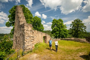 Zwei Wanderer laufen auf einer Wiese neben den Überresten einer alten Burg unter blauem Himmel und umgeben von eingien Bäumen. 