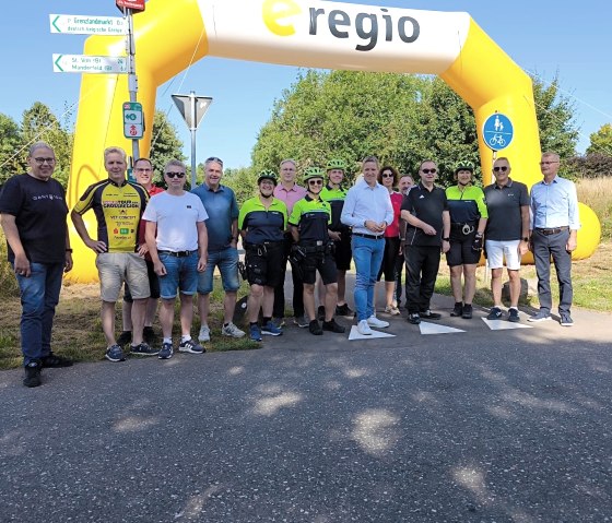 Zahlreiche Personen, welche teilsweise sportlicher Radfahrkleidung tragen, stehen auf dem Radweg unter einem großen Luftbogen des Sponsors e-regio.