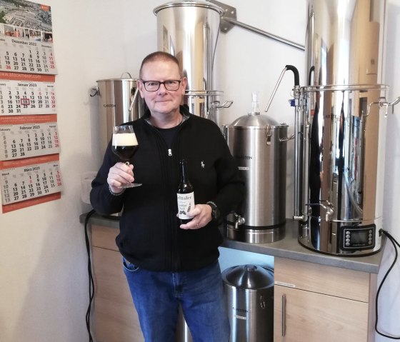 Ein Mann steht vor einer Brauanlage und h&auml;lt eine Flasche Bier und ein gef&uuml;lltes Glas hoch. 