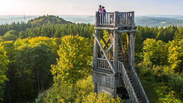Holzaussichtsturm im Wald mit zwei Personen, umgeben von grünen Bäumen und Hügeln im Hintergrund.