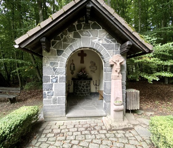 Front view of the small Arnulphus Chapel with a small altar and painted walls inside. A paved path leads to the chapel. Behind it is a dense deciduous forest., &copy; Touristik GmbH Gerolsteiner Land