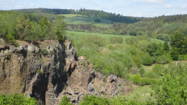 Felsen und grüne Hügel im Vulkangarten Steffeln, umgeben von Bäumen und Windrädern am Horizont.