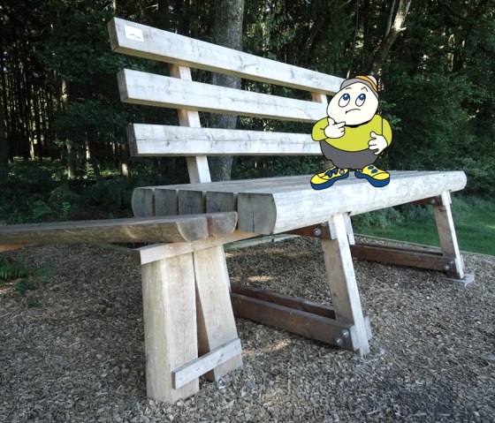 Cartoonfigur Willi Basalt mit grünem Shirt, grauer Hose und gelben Schuhen steht auf einer überdimensionalen XXL Bank in einem Wald.