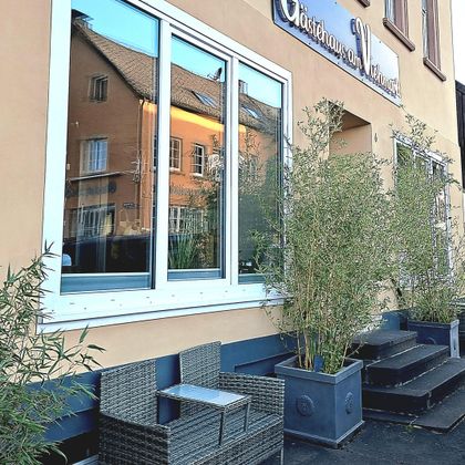 Ein gemütliches Gasthaus mit großen Fenstern und stilvollen Pflanzen. Vor dem Eingang stehen moderne Sitzmöbel auf einer kleinen Terrasse.