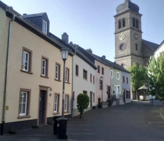 Eine malerische Stra&szlig;e von Hillesheim mit pastellfarbenen H&auml;usern und der Pfarrkirche mit Turm im Hintergrund. Der Himmel ist klar und blau., &copy; Albert Junk