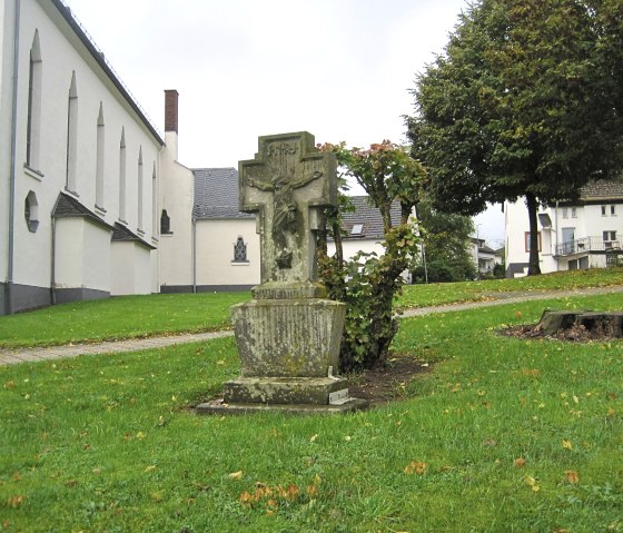Ein Steinkreuz steht auf einer grünen Wiese vor einer weißen Kirche. Im Hintergrund sind Bäume und Häuser zu sehen., © Touristik GmbH Gerolsteiner Land, Ute Klinkhammer