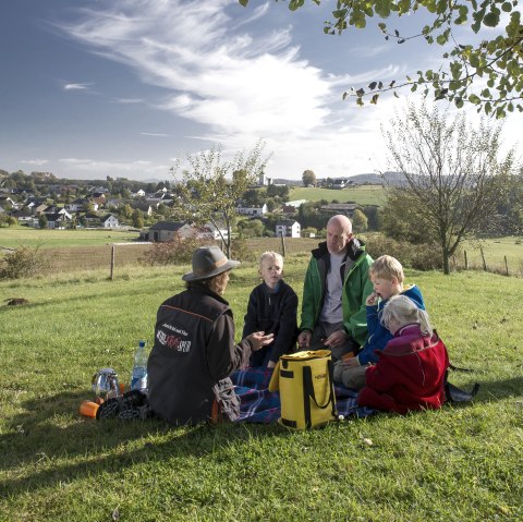 Two adults and two children sit on a blanket in a spacious meadow next to a tree. They have provisions in rucksacks.