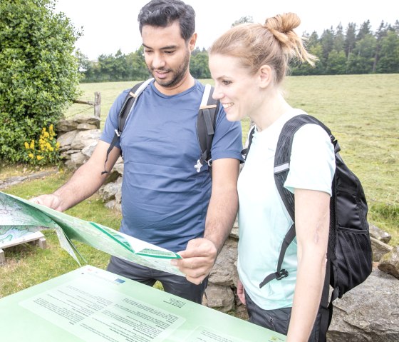 Zwei Personen mit Rucksäcken betrachten eine Wanderkarte vor einer grünen Wiese und Bäumen., © Eifel Tourismus GmbH, AR-shapefruit AG