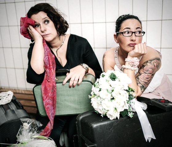 Two women in smart clothes sit next to each other with sad faces, their packed suitcases in front of them.