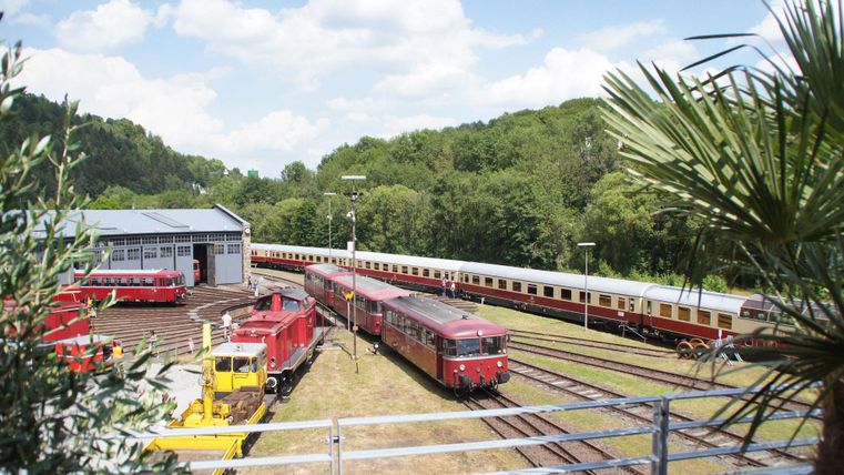 Ein Bahnhof mit mehreren Zügen und einem Reparaturgebäude. Im Hintergrund sind Bäume und ein blauer Himmel zu sehen.