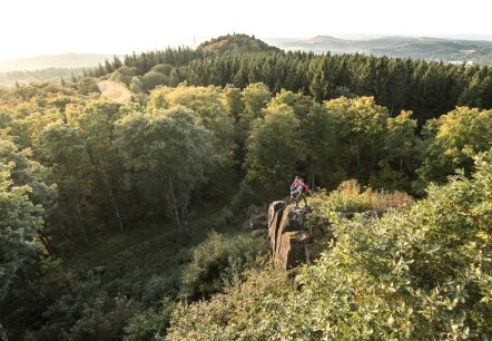 Two people are standing on a rock surrounded by a dense forest. Hills and a wide view over the landscape can be seen in the background., © Eifel Tourismus GmbH, D. Ketz