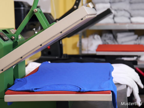 Ein blaues T-Shirt liegt in einer Stoffpresse. Im Hintergrund sieht man ein Reagl mit gefalteten weißen T-Shirts.