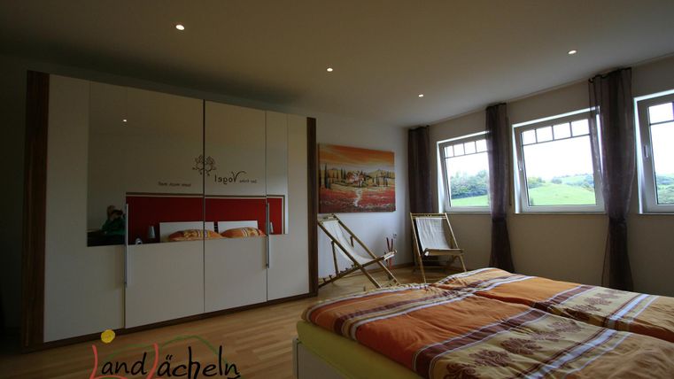 Ein modernes Schlafzimmer mit einem Doppelbett und hellen Holzböden. Große Fenster lassen viel Tageslicht herein und bieten einen Blick auf die Landschaft.