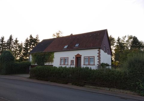 Une maison simple à deux étages avec un toit à deux versants rouge et des murs blancs. Elle est entourée d'arbres et d'une haute haie, dans un environnement rural paisible.
