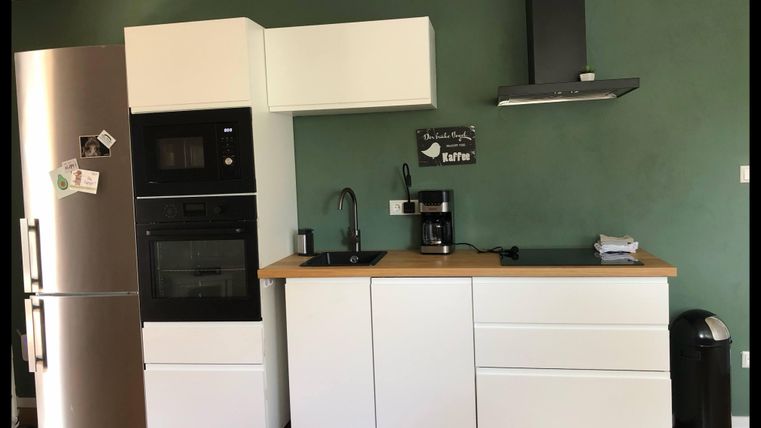 Eine moderne Küche mit weißen Möbeln und einer Holzarbeitsfläche. Im Hintergrund befindet sich eine grüne Wand und Küchengeräte wie ein Kühlschrank und eine Kaffeemaschine.